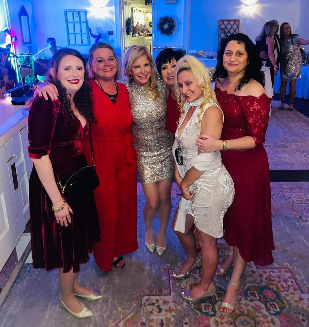 Six women dressed in elegant evening attire, smiling and posing together at a party or celebration in a decorated venue with blue lighting.