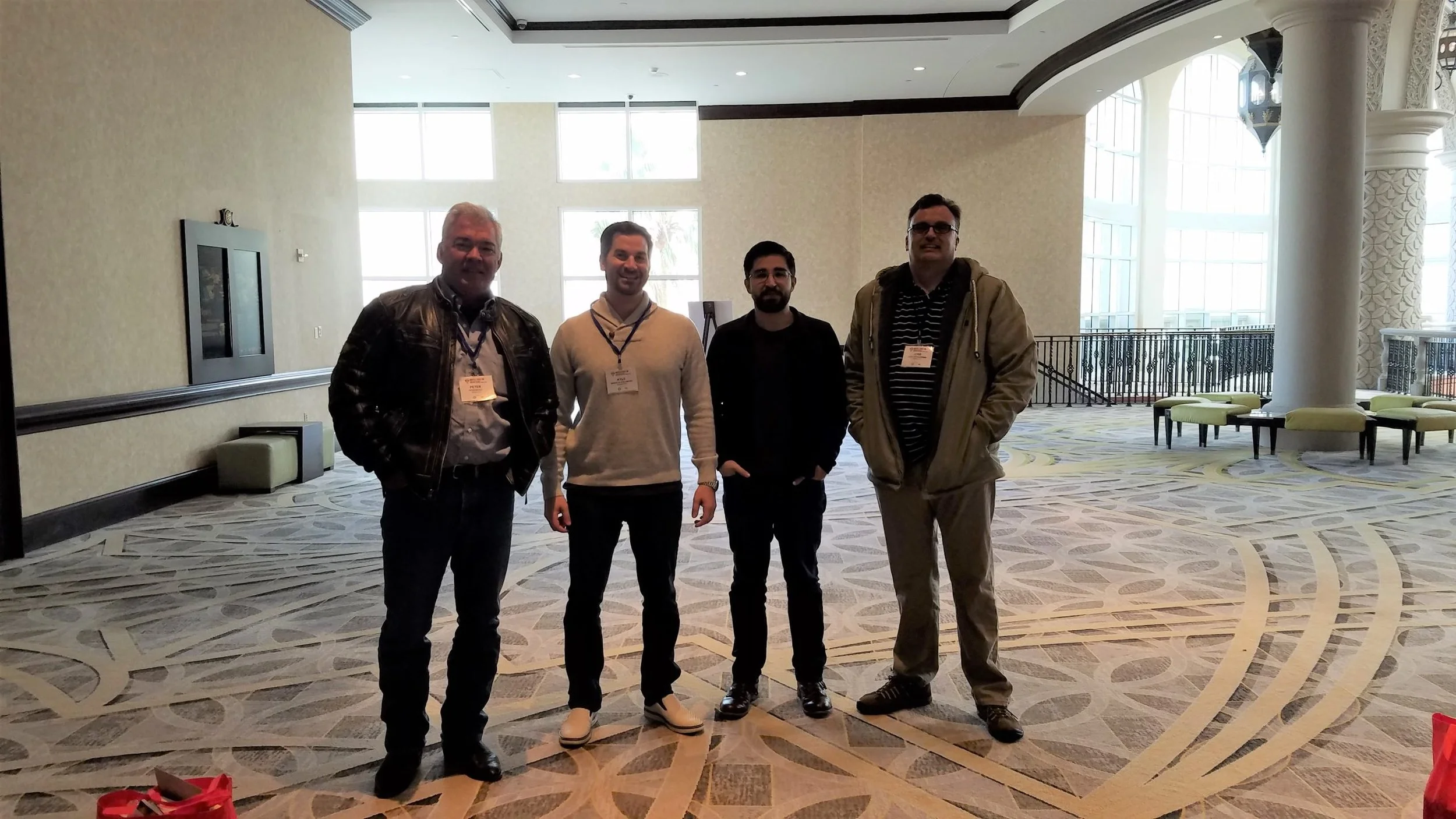 Four men standing together in a hotel lobby with large windows, patterned carpet, and elegant decor.