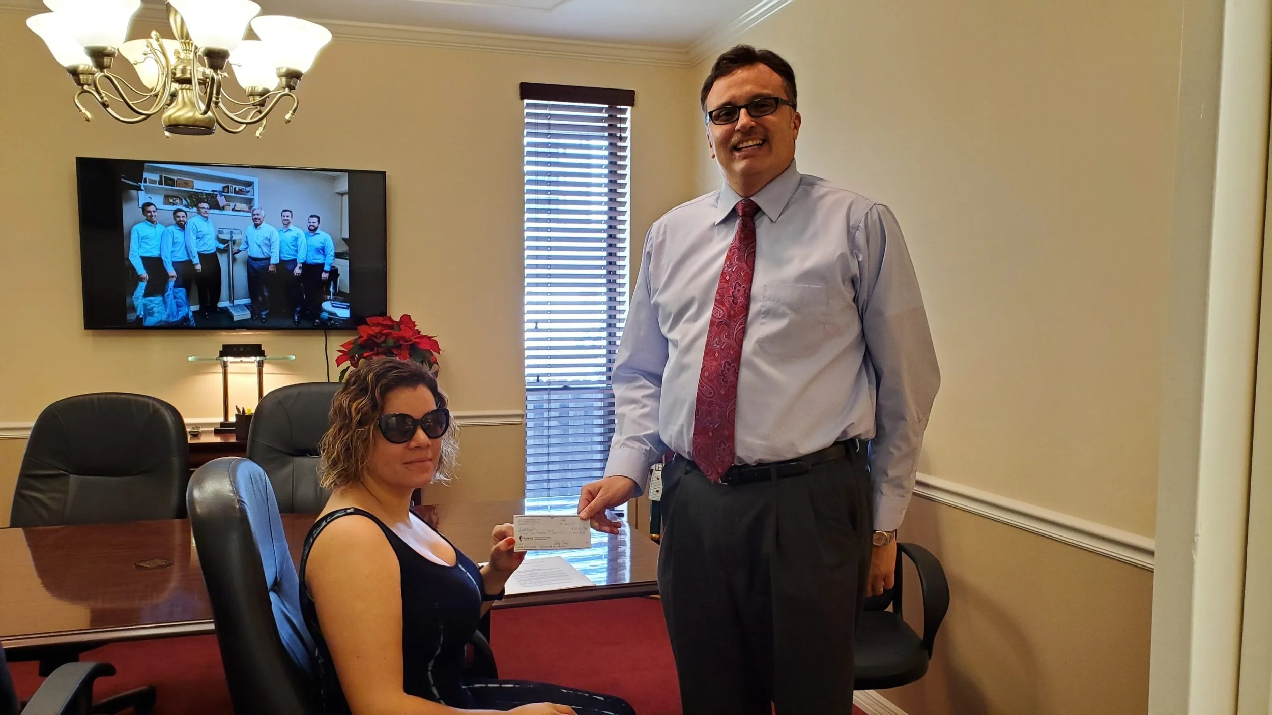 A woman seated at a conference table receiving a check from a man in a dress shirt and tie in a professional office setting with a television displaying a group photo in the background.