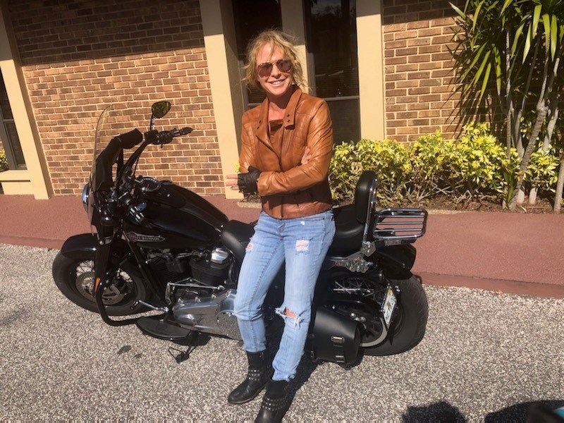 A woman wearing a brown leather jacket, ripped jeans, and black boots standing next to a black motorcycle on a sidewalk outside a brick building with yellow-green plants.