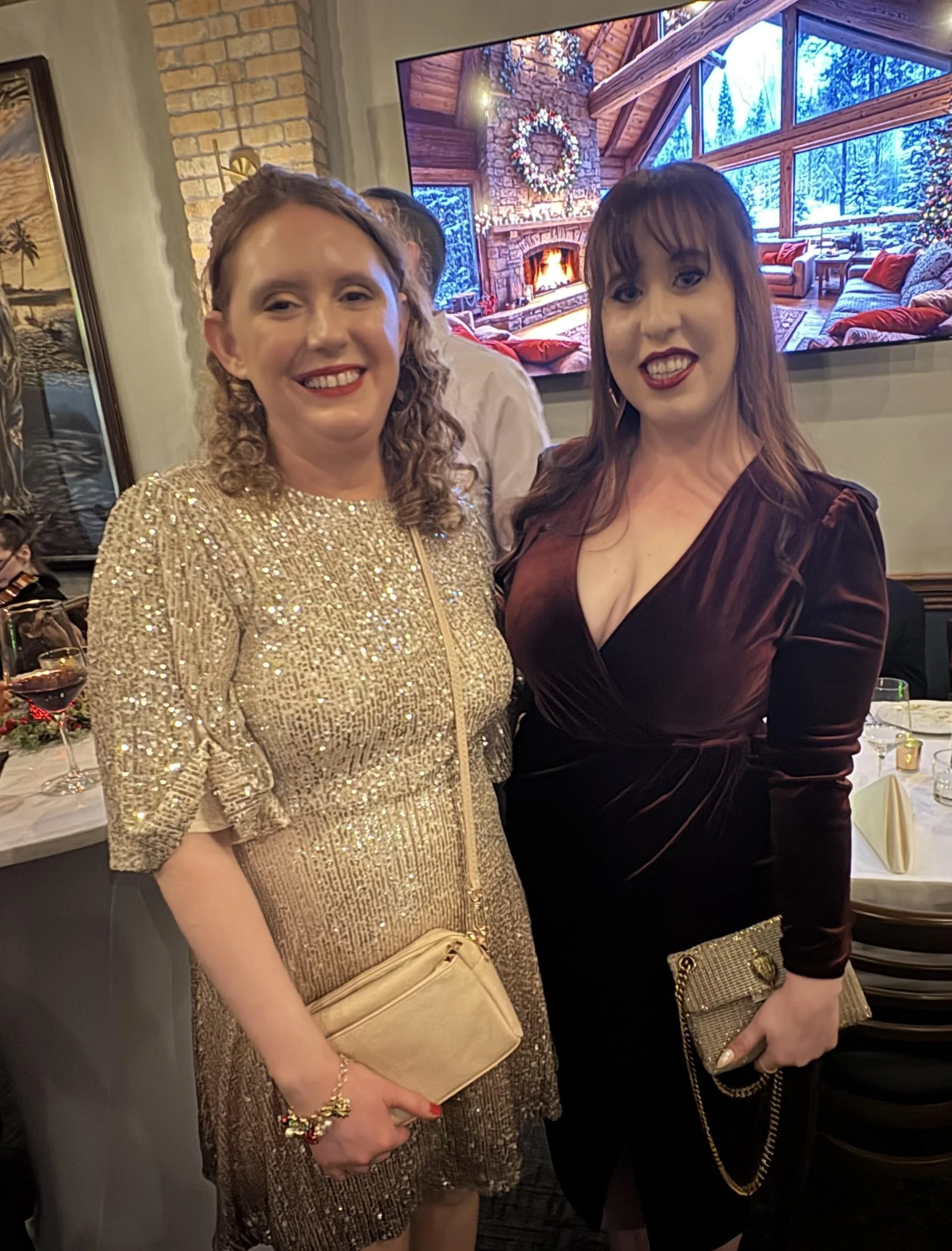 Two women smiling and posing at a celebration or party, with festive table settings and a large television displaying a cozy fireplace scene in the background.