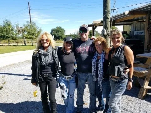 Group of five people posing outdoors on a sunny day, standing on gravel near a wooden structure and utility pole.