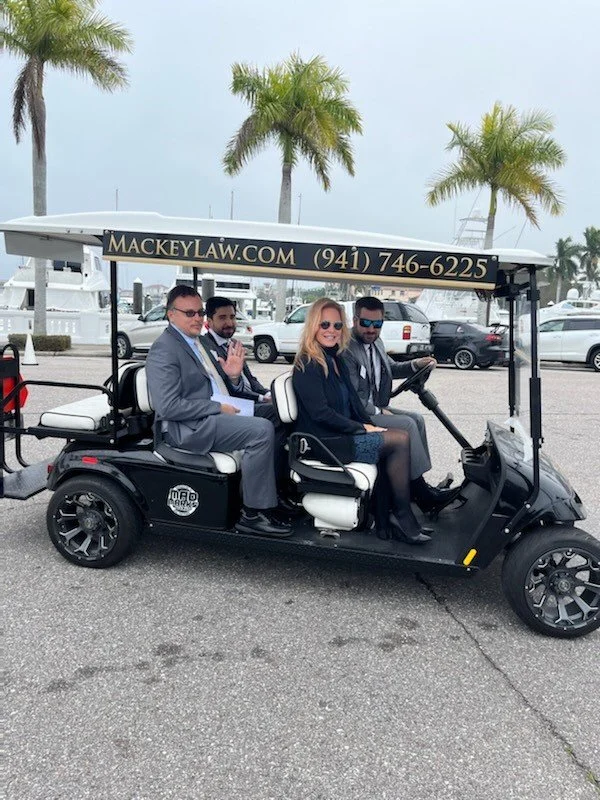 Four people in formal attire sitting on a black golf cart with a sign for Mackey Law, parked in a lot with palm trees and other vehicles in the background.