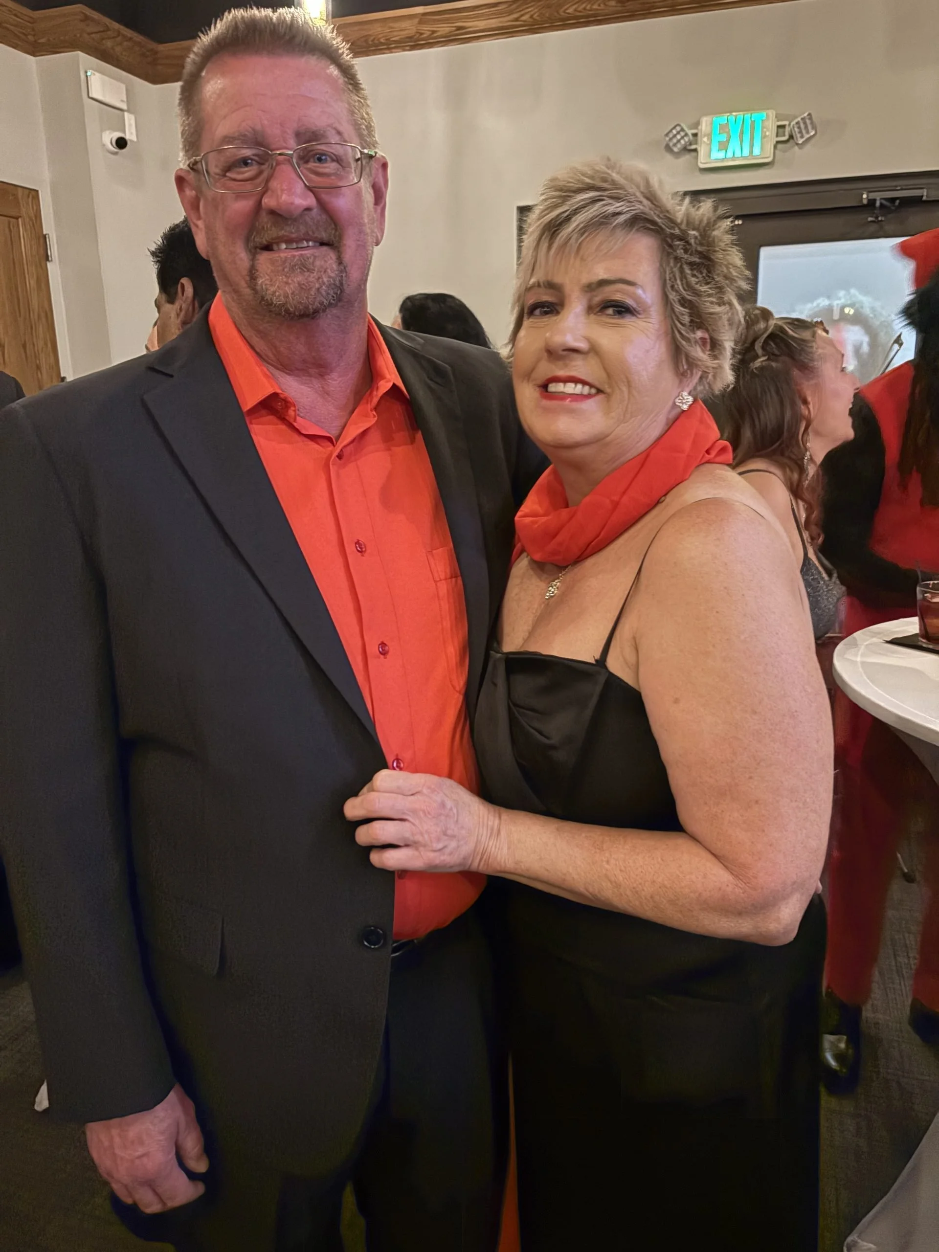 A smiling man and woman are standing close together, holding hands at an indoor event. The man is wearing glasses, a black suit jacket, and an orange shirt. The woman has short, blonde hair and is dressed in a black dress with a red scarf around her 