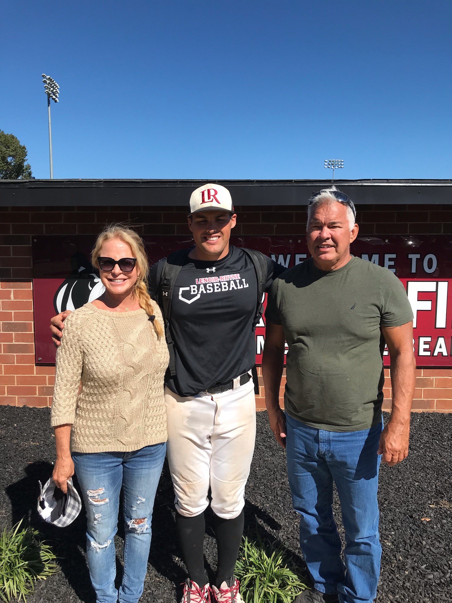 Three people posing in front of a sign at a sports stadium, including a woman with sunglasses and a beige sweater, a baseball player in uniform, and a man in a green shirt and jeans.