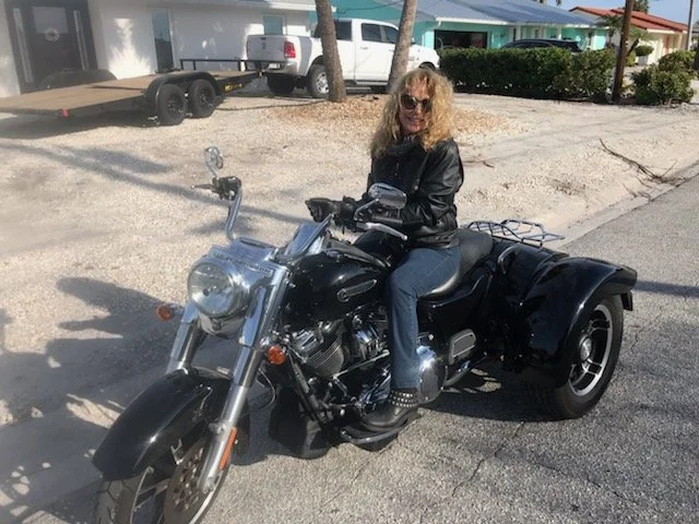 A woman sitting on a black motorcycle with a sidecar, wearing sunglasses and a black jacket, on a sunny street with houses and parked cars in the background.