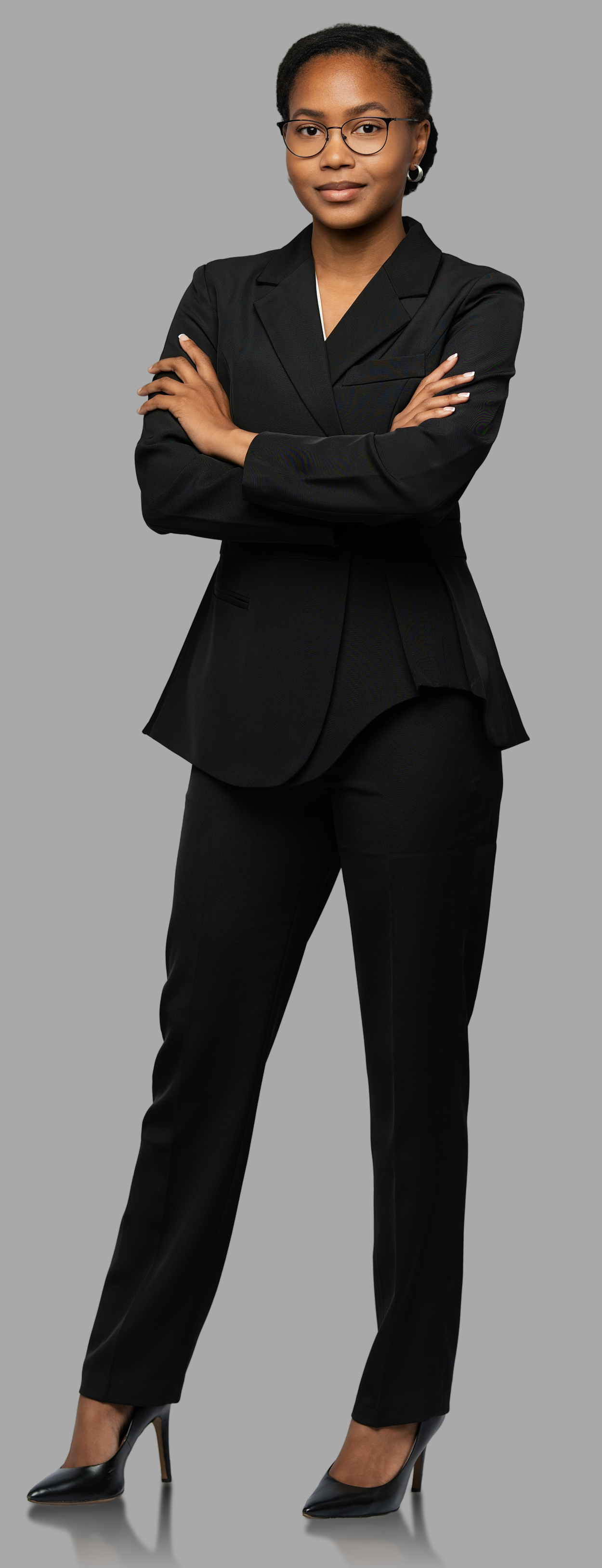 A confident African American woman in a black business suit with crossed arms, wearing glasses, standing against a plain gray background.