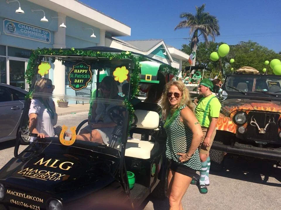 People celebrating St. Patrick's Day in a parade, with a golf cart decorated with green shamrocks and a sign that says "Happy St. Patrick's Day!" along with a group of vehicles, including a fire truck and a guitar-shaped float, in a sunny outdoor set