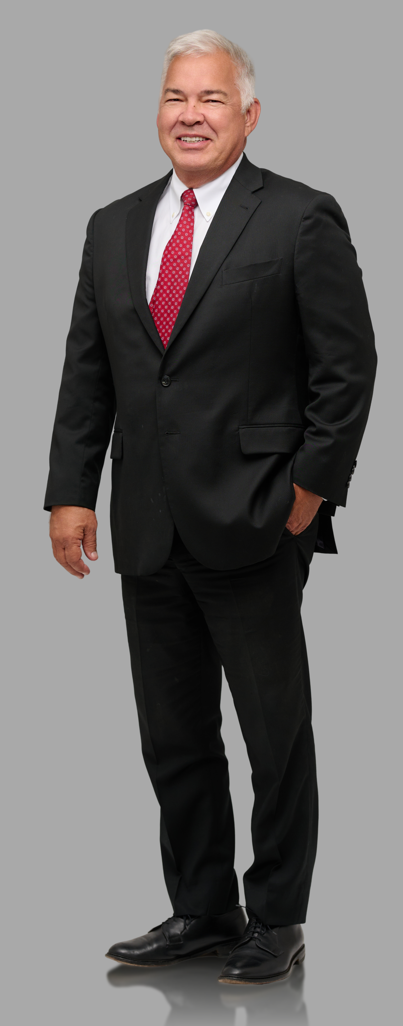 A smiling middle-aged man with gray hair, dressed in a black suit, white shirt, and red polka-dot tie, standing against a plain gray background.