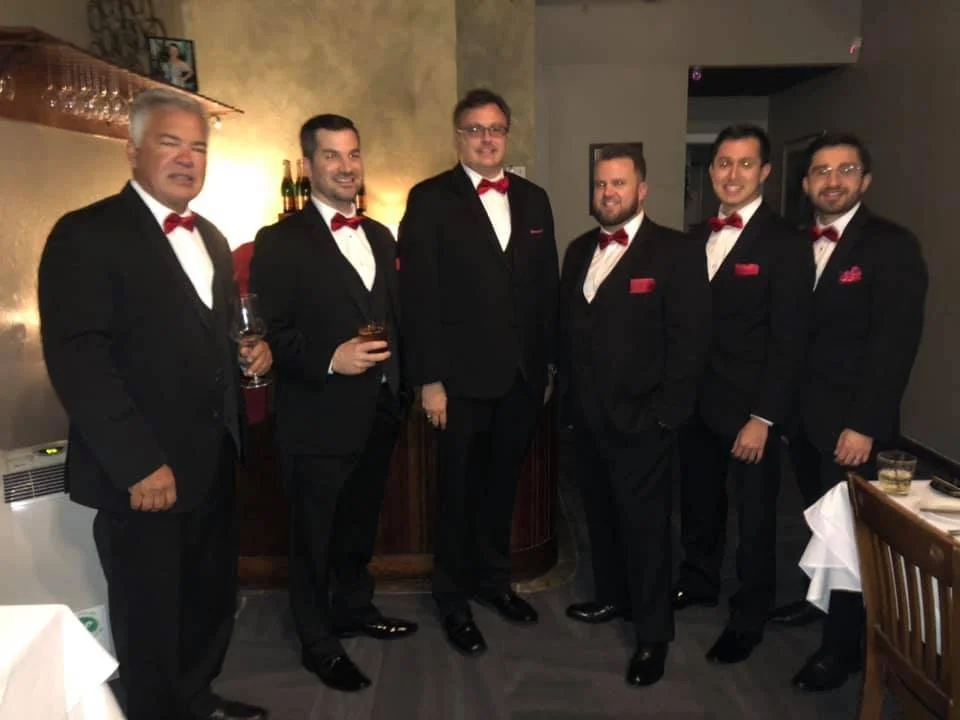 Seven men dressed in formal black tuxedos with red bow ties and pocket squares, standing together in a restaurant or banquet setting.