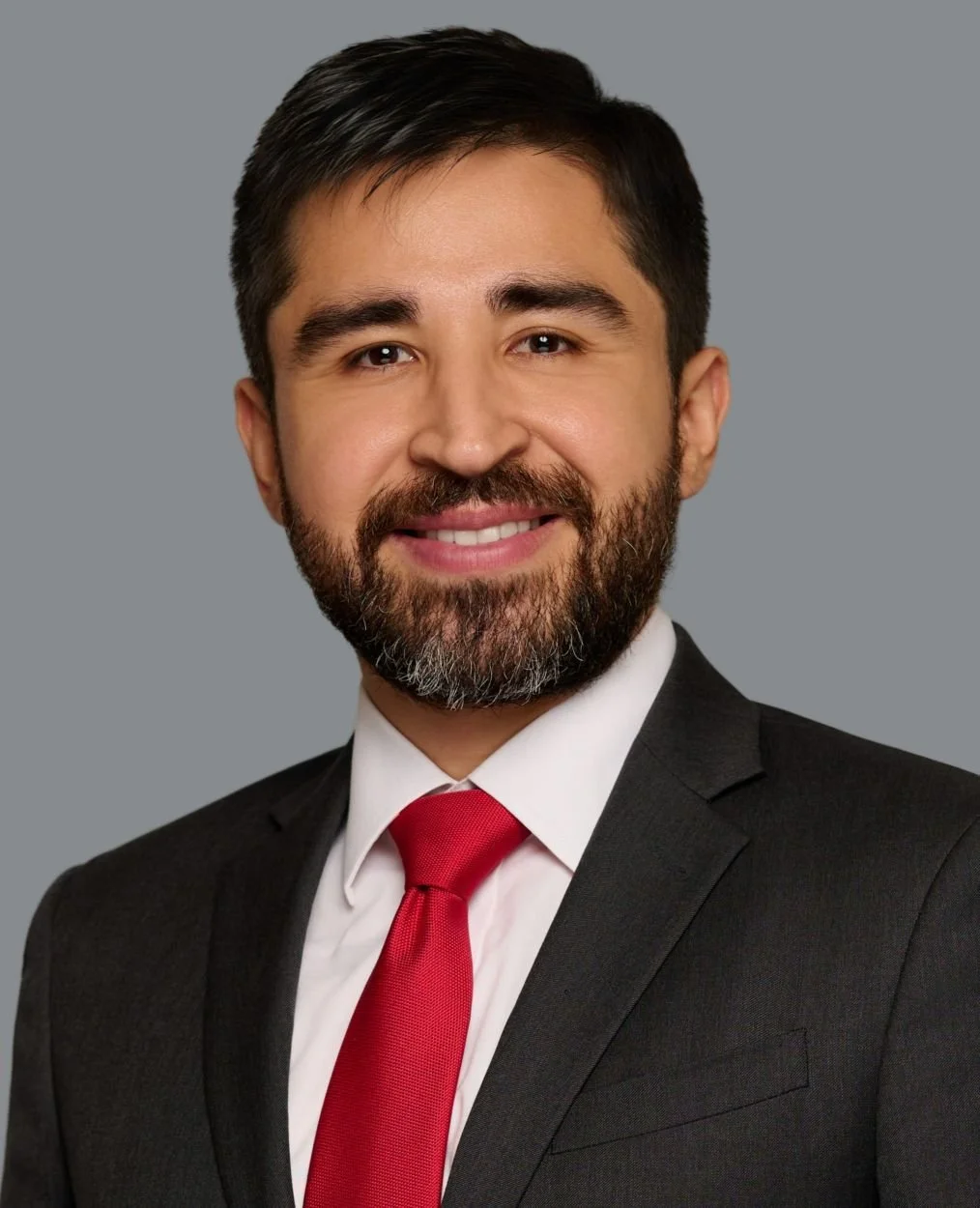 Headshot of a man with dark hair and beard, wearing a dark suit, white shirt, and red tie, smiling against a gray background.