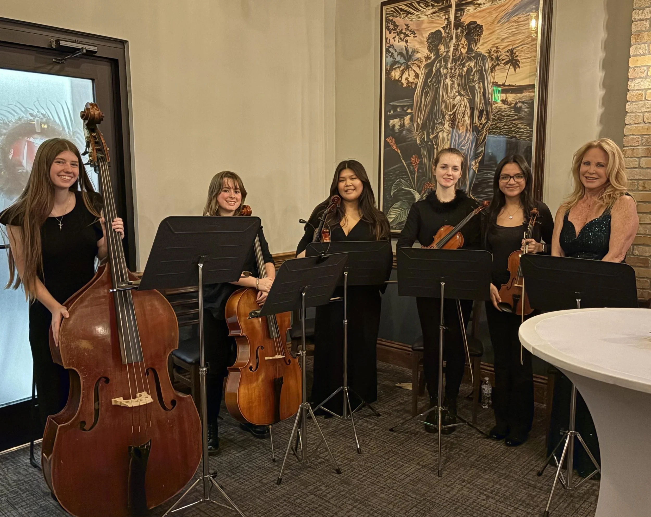 Group of five women musicians holding string instruments, standing behind music stands in a room with artwork on the wall and a brick column.