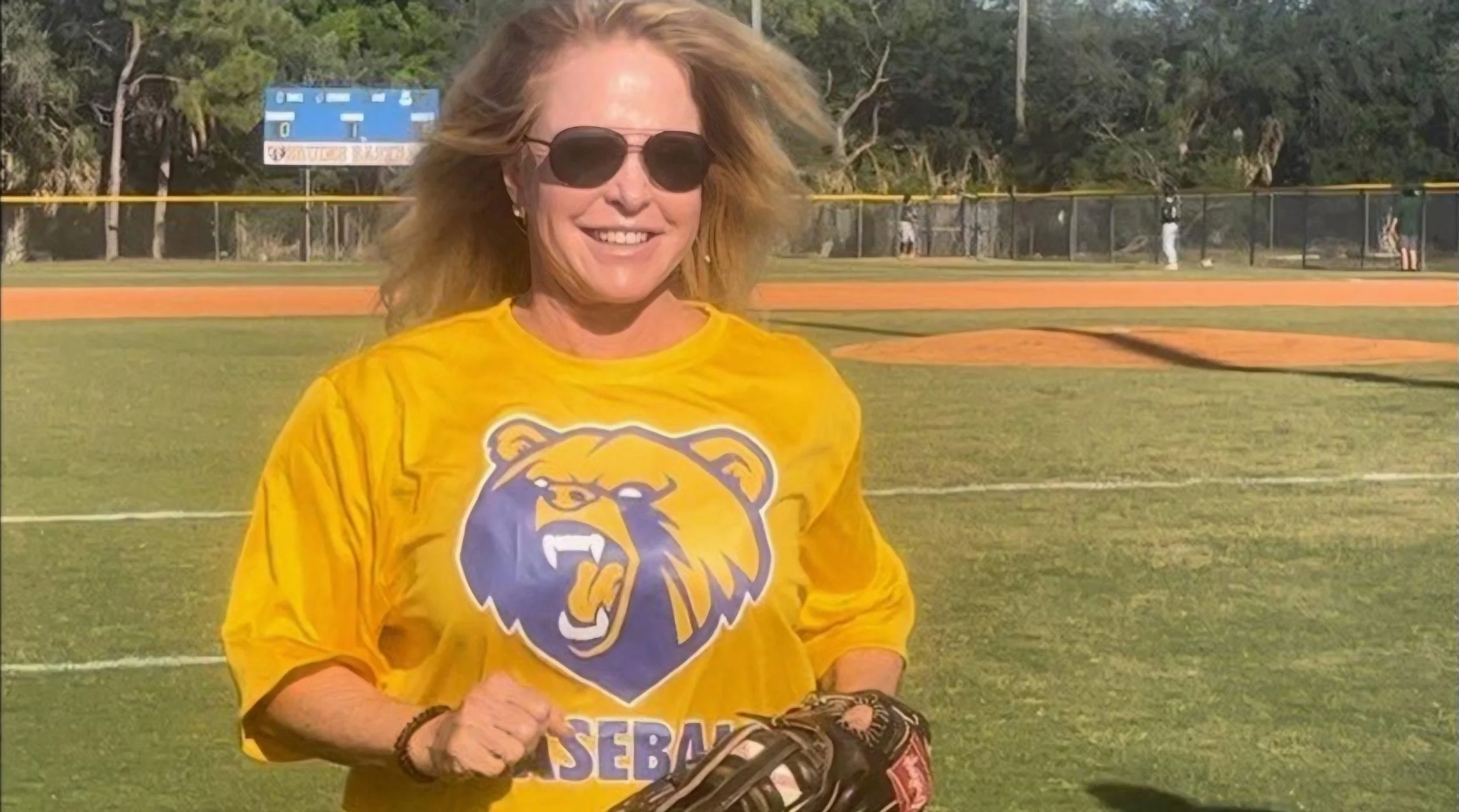 A woman with red hair wearing sunglasses and a yellow shirt with a bear logo, standing on a baseball field, smiling at the camera.
