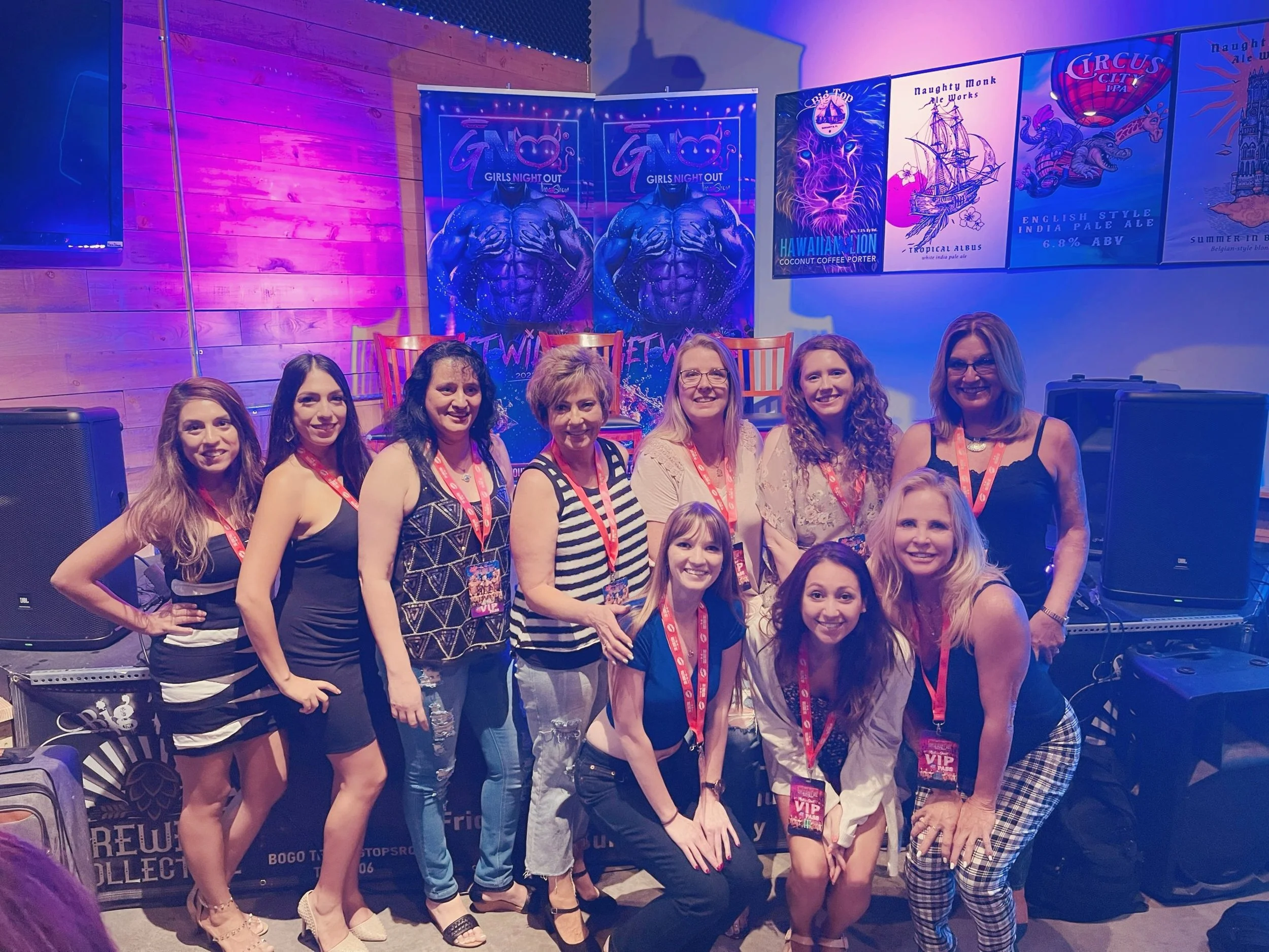 A group of women and one girl posing together at an indoor event with colorful banners and posters on the wall behind them, including promotional posters for gin and beer brands, and a purple lit background.