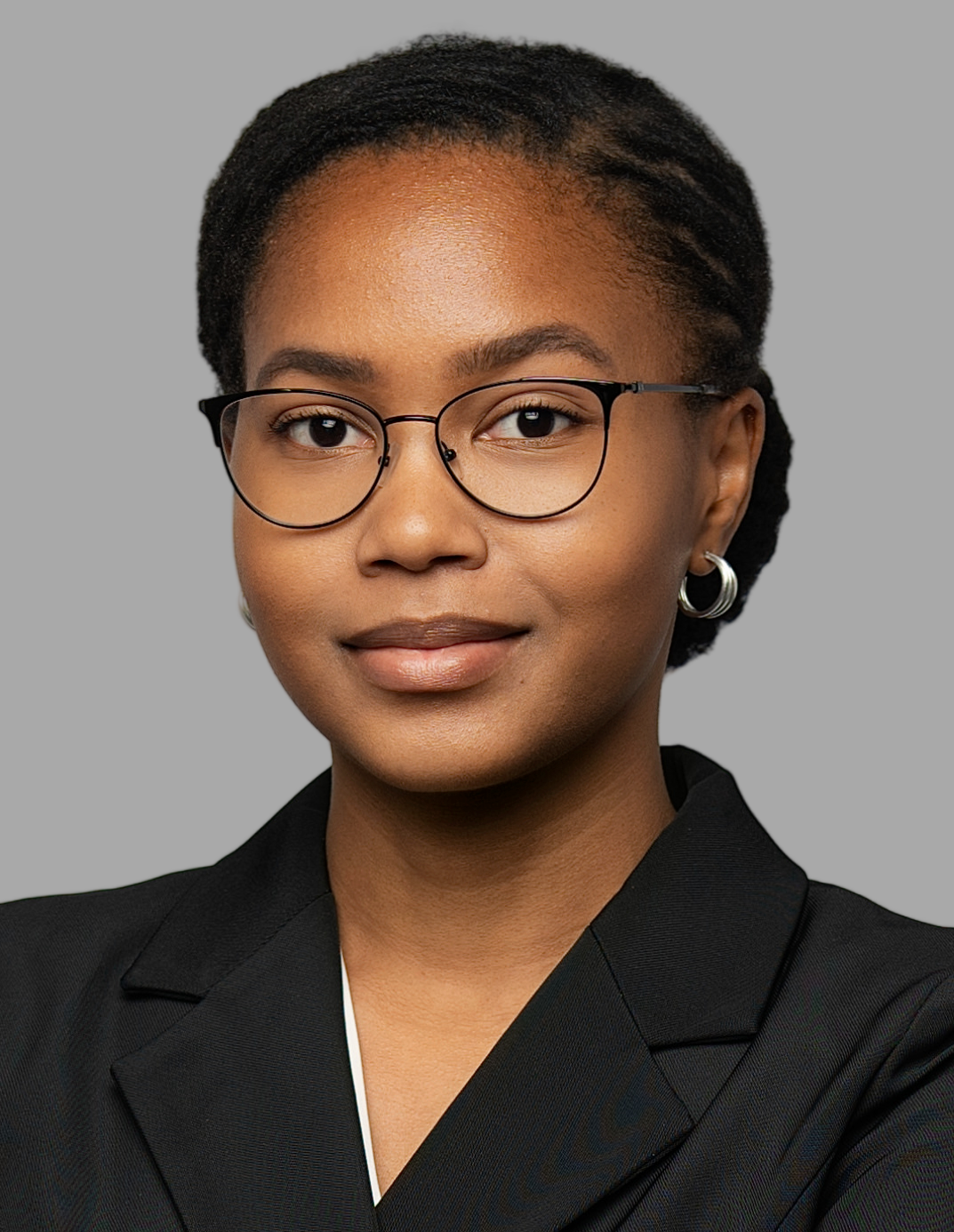 A professional woman with glasses and hoop earrings, wearing a black blazer, smiling with a neutral gray background.