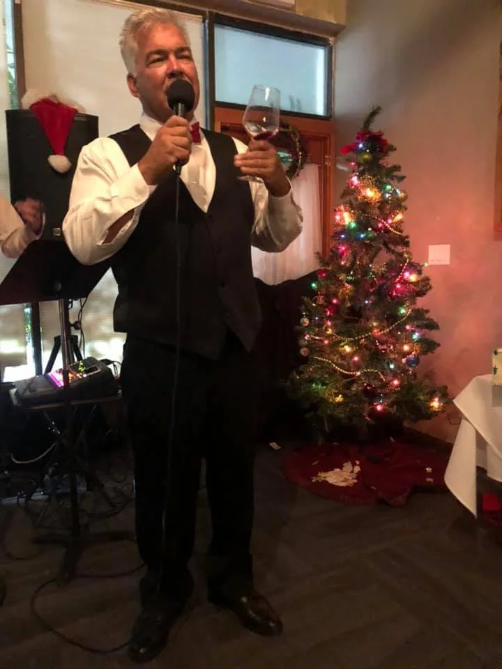 A man in a black vest, white shirt, and red bow tie, holding a microphone and a glass of wine, standing next to a decorated Christmas tree with colorful lights and ornaments.