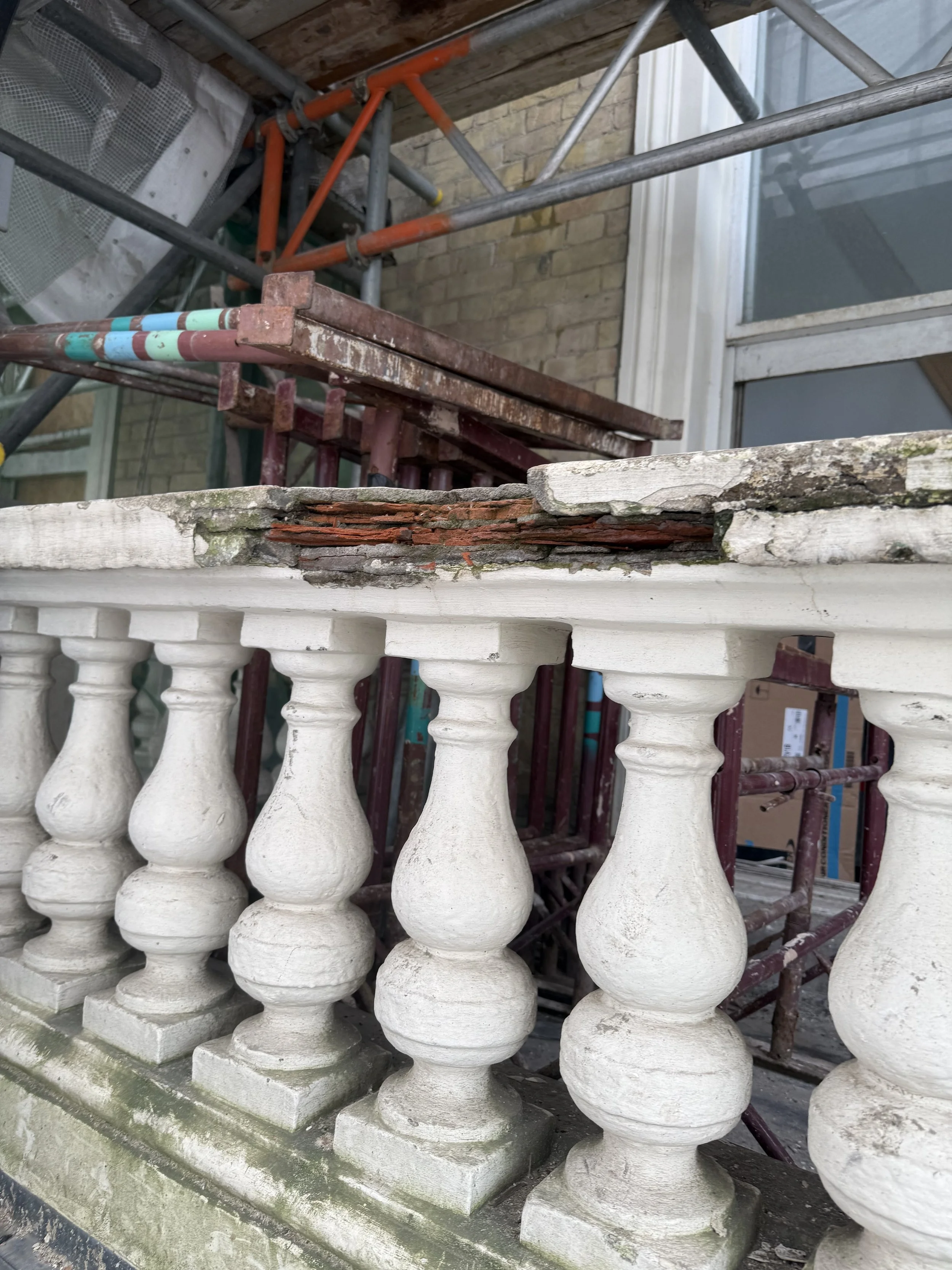 Close-up of a weathered white concrete balustrade with chipped and cracked decorative balusters, set against scaffolding and a brick wall.