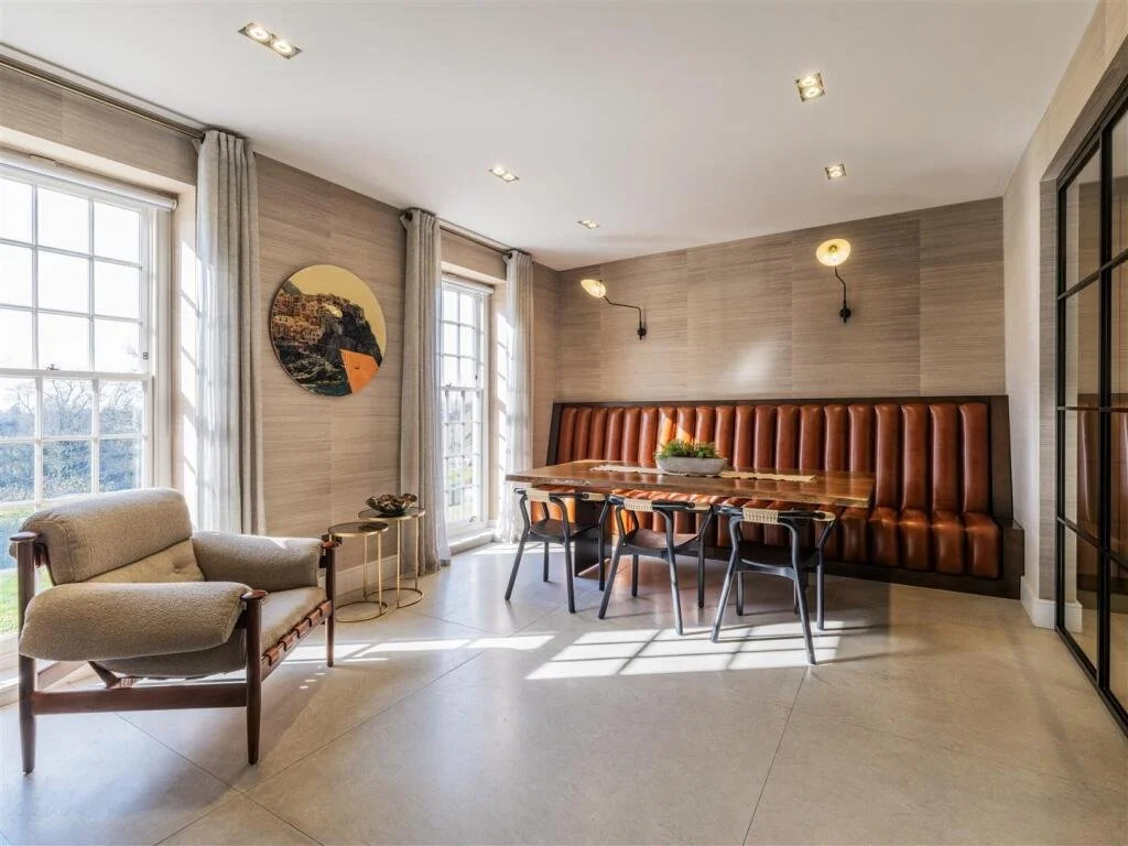 Bright living room with large windows, beige curtains, a beige armchair, a round side table, wall art, a long wooden dining table, a leather-upholstered bench, and black chairs, all illuminated by wall-mounted lights.