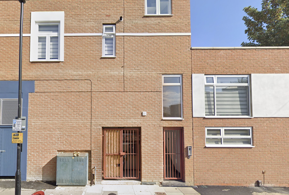 Facade of a modern brick building with multiple windows, two red metal gates, a utility box, a parking sign, and a lamppost.