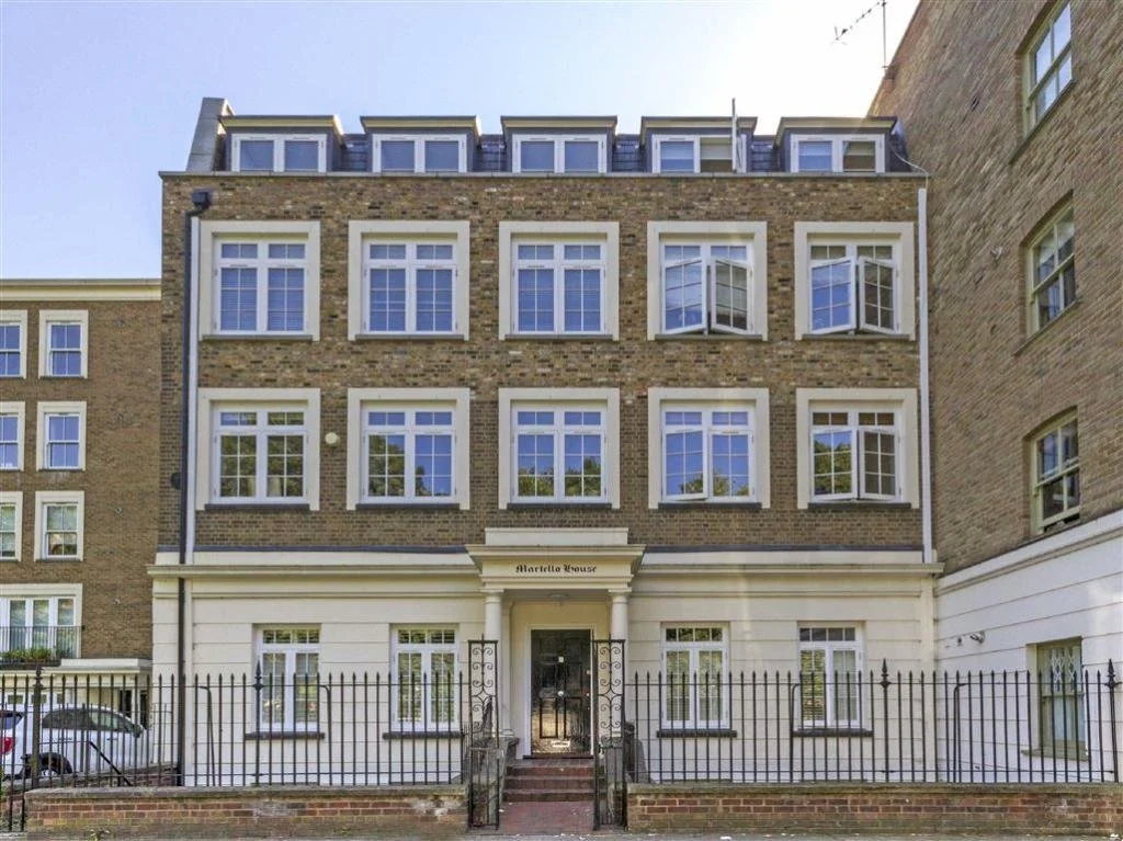 A multi-story brick apartment building with white window frames and a white entrance wall, gated by a black metal fence, with a small garden area and parked cars nearby.