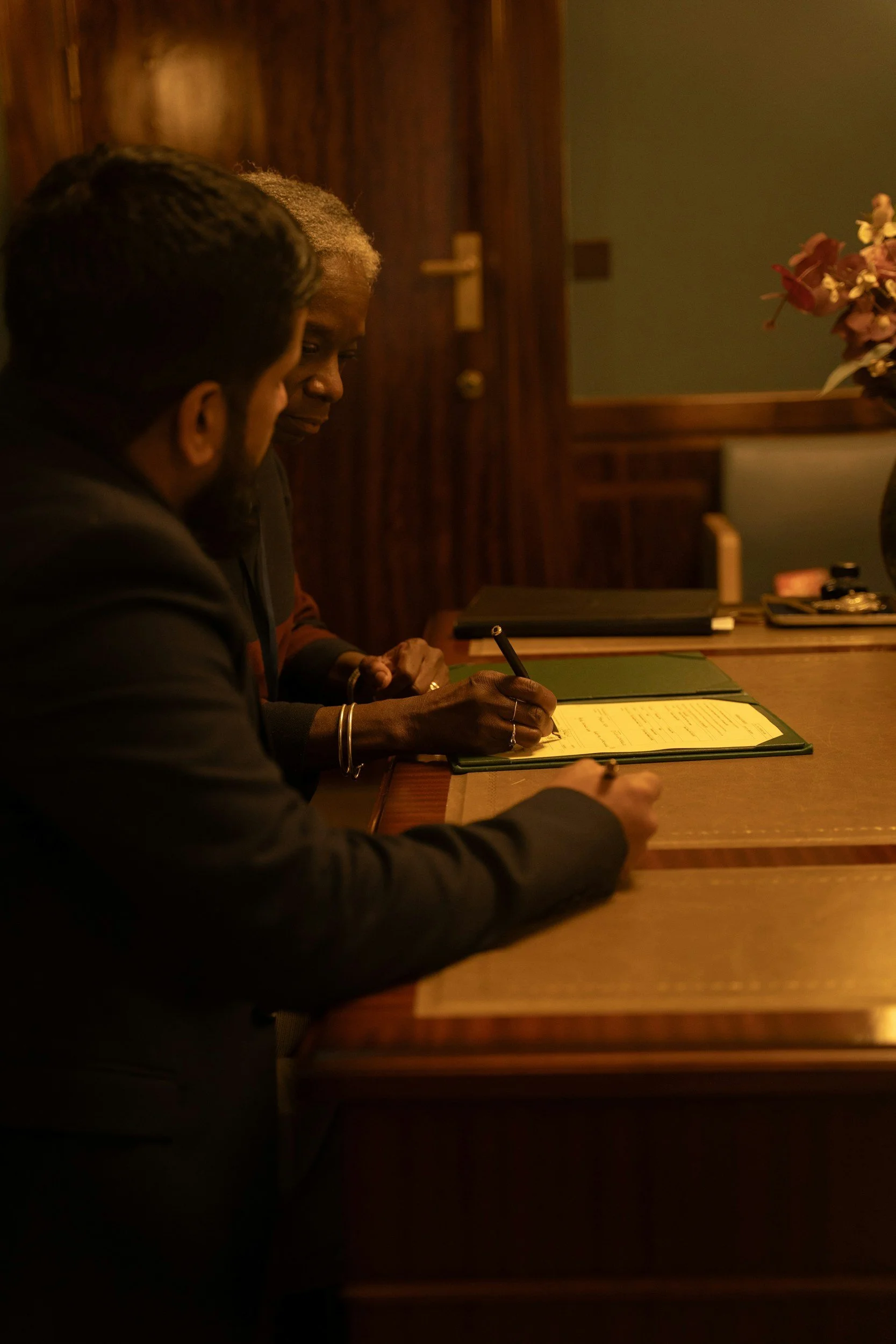 Two people sitting at a wooden table, writing in a yellow notepad with a black pen, in a warmly lit room with wooden paneling.