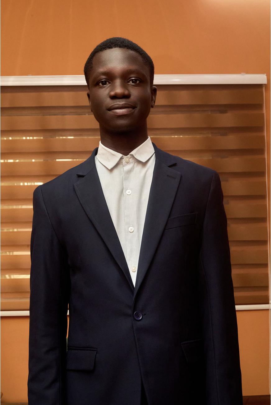 A young man in a dark suit and white shirt standing indoors in front of wooden blinds.