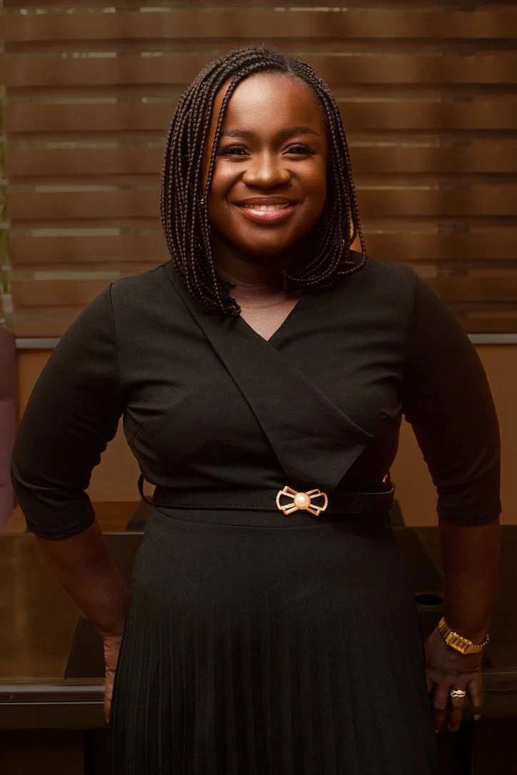 A woman with braided hair smiling and wearing a black dress with a decorative belt, standing in front of a wooden wall.