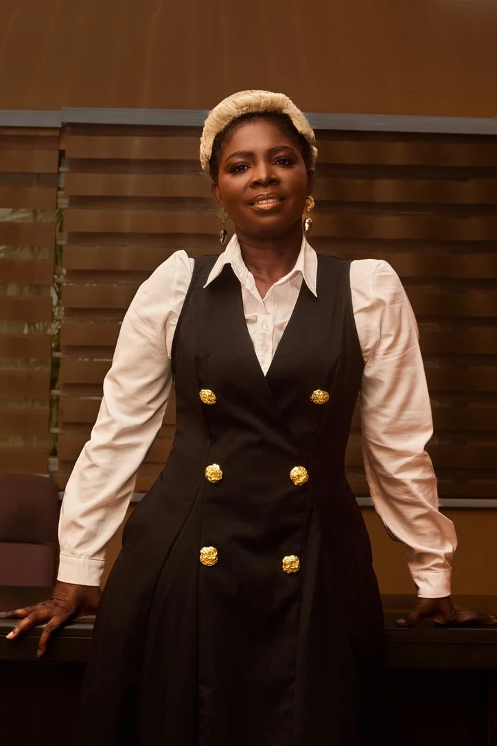 A woman standing in front of a wooden-paneled wall, wearing a white blouse with puffed sleeves, a black dress with gold buttons, gold earrings, and a beige headband.