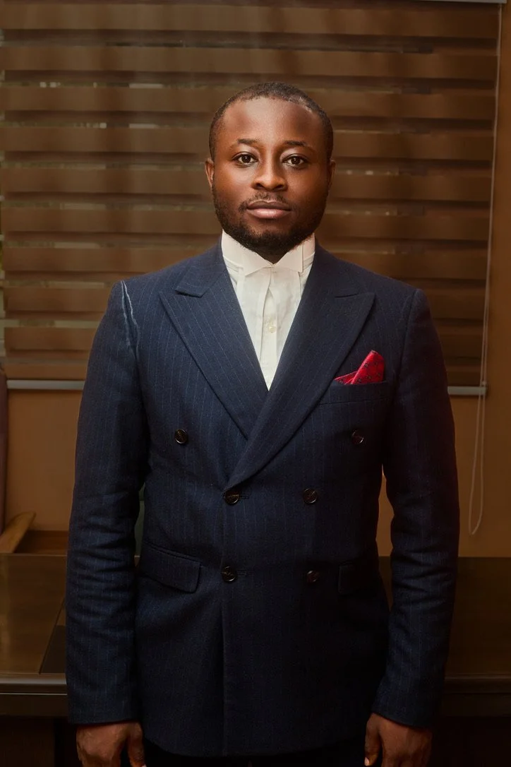 A man dressed in a dark, double-breasted suit with a white shirt and a red pocket square, standing indoors in front of wooden blinds.