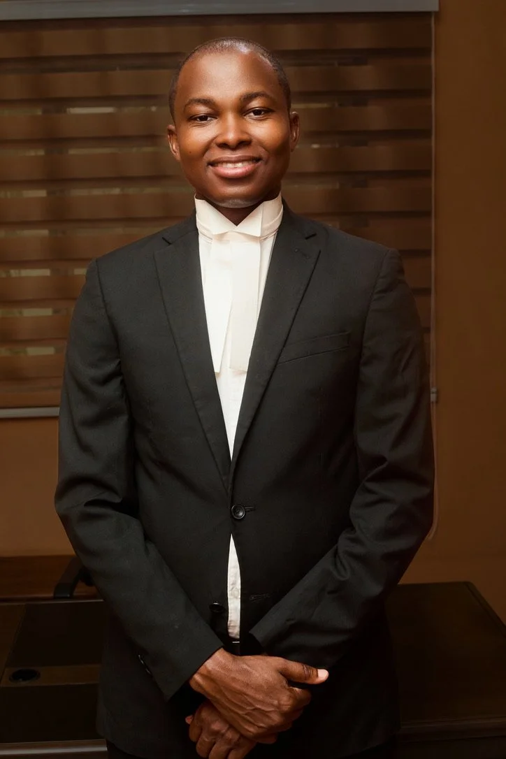 A person in a black suit and white shirt standing indoors, smiling at the camera with hands clasped in front.