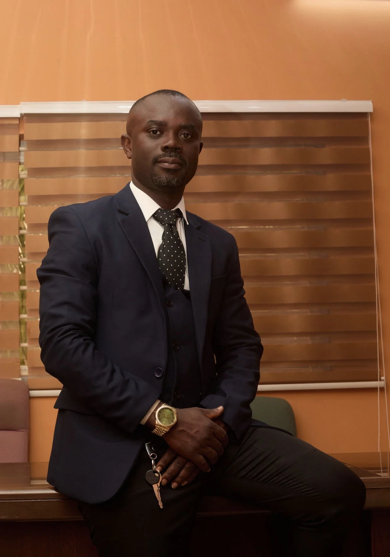A man in a navy suit and polka dot tie sitting in front of wooden blinds, with a gold watch and a set of keys in his hand.