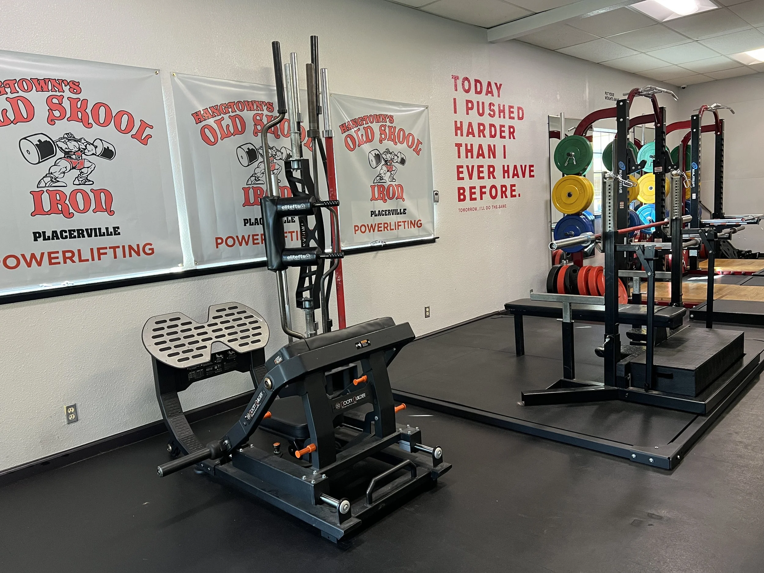 Inside a gym, with banners on the wall that read 'HANGTOWN'S OLD SCHOOL IRON PLACERVILLE POWERLIFTING' and motivational quote 'TODAY I PUSHED HARDER THAN I EVER HAVE BEFORE.' Weightlifting equipment, including a squat rack with weights and a seated c