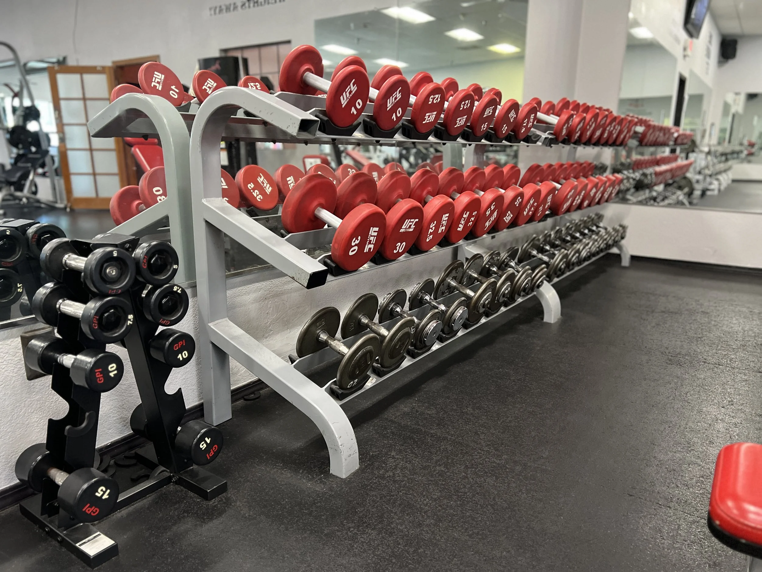 A set of red and black dumbbells organized on a multi-tiered rack at a gym. The background shows a mirror, some workout equipment, and a door with a window.