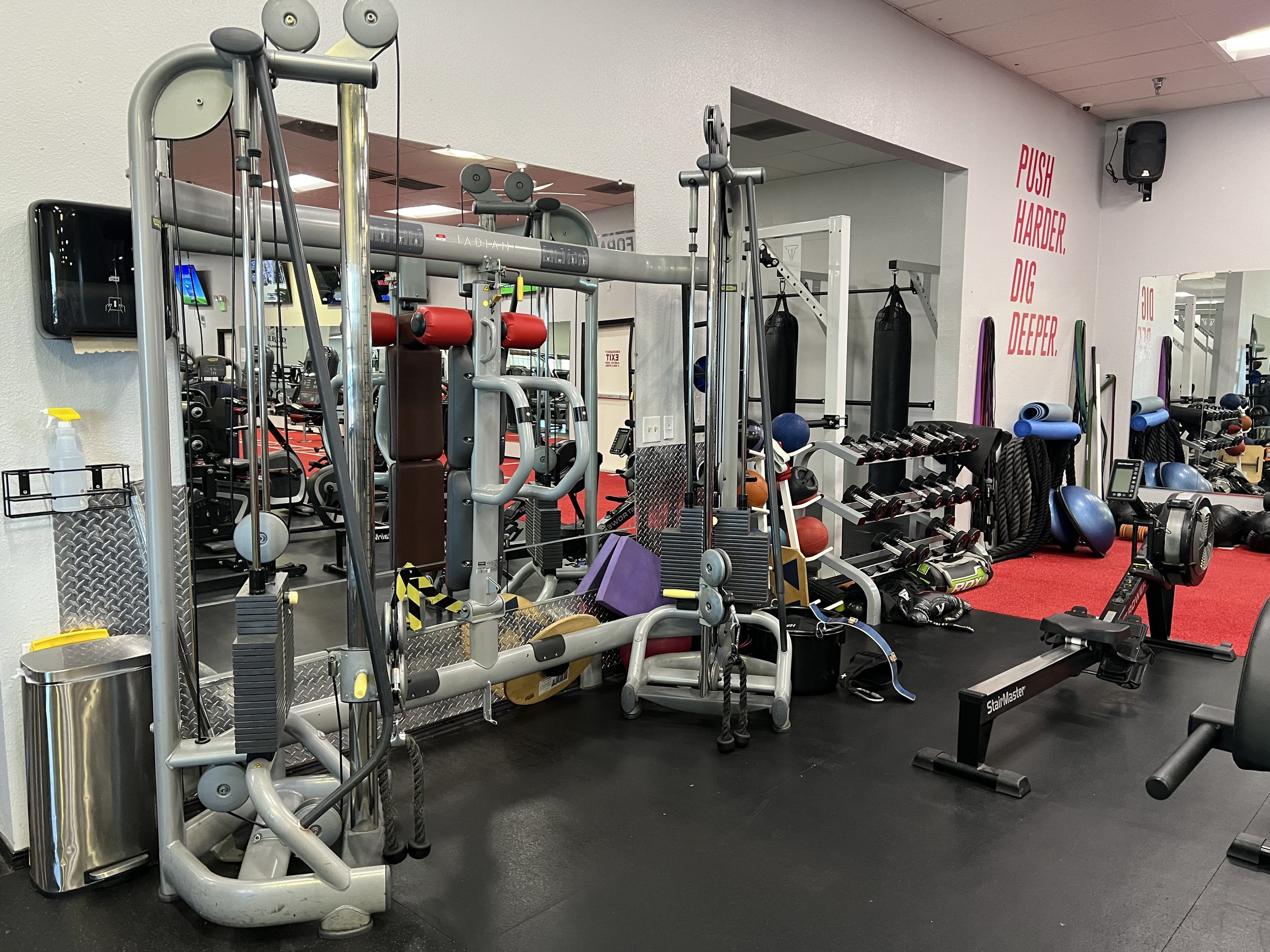 Gym equipment including weights, punching bags, and workout machines in a fitness center with a motivational sign that reads 'Push Harder, Dig Deeper.'