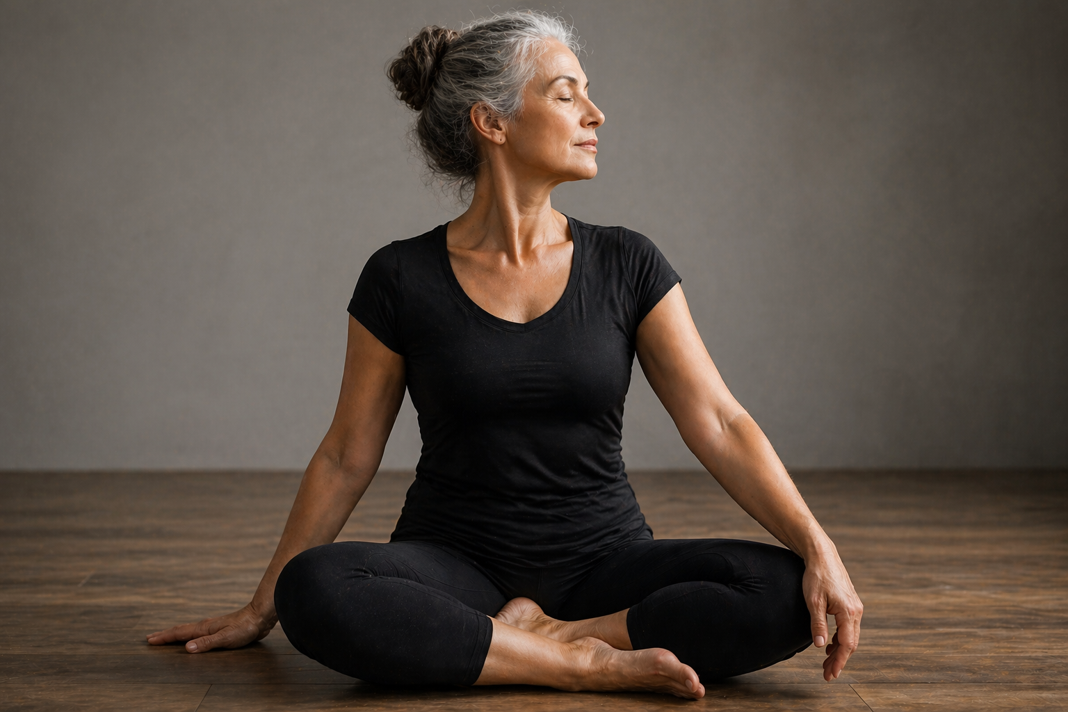 An elderly woman with gray hair tied back practicing yoga, seated cross-legged on a wooden floor with a plain gray background, eyes closed, in a meditative pose.