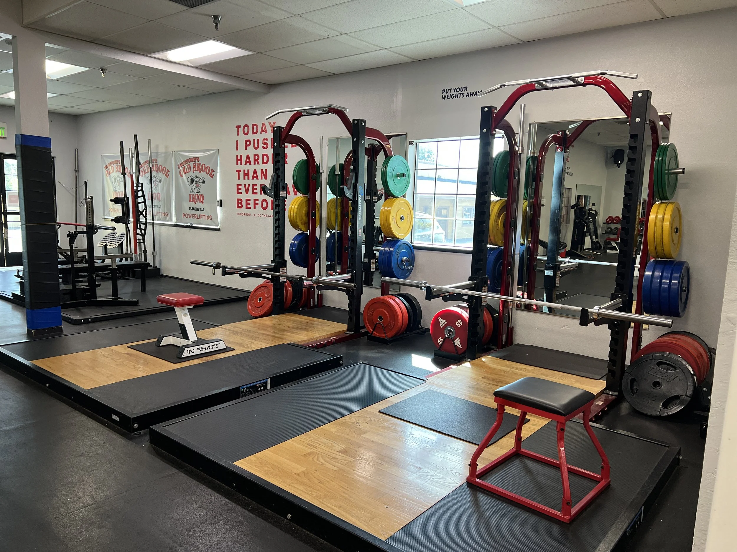 Interior of a gym with weightlifting equipment, including squat racks with weight plates, a bench and various exercise machines. The gym has a motivational quote on the wall reading 'Put your weights away,' and a large window letting in natural light