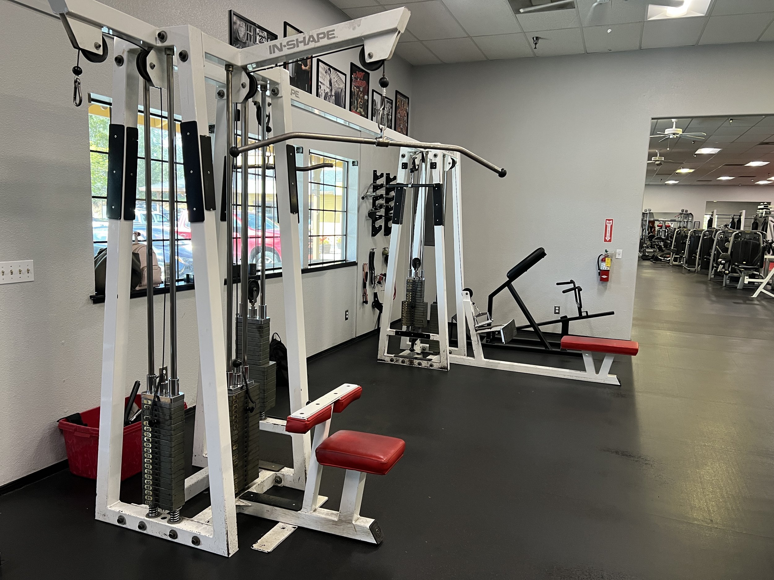 An empty gym workout area with weightlifting equipment, including a machine with weights, a leg press, and other exercise equipment, with large windows and framed photos on the wall.