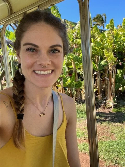 A young woman with light skin, a braid, and a septum piercing, smiling at the camera, wearing a yellow top, standing outside near banana plants and a bright blue sky.