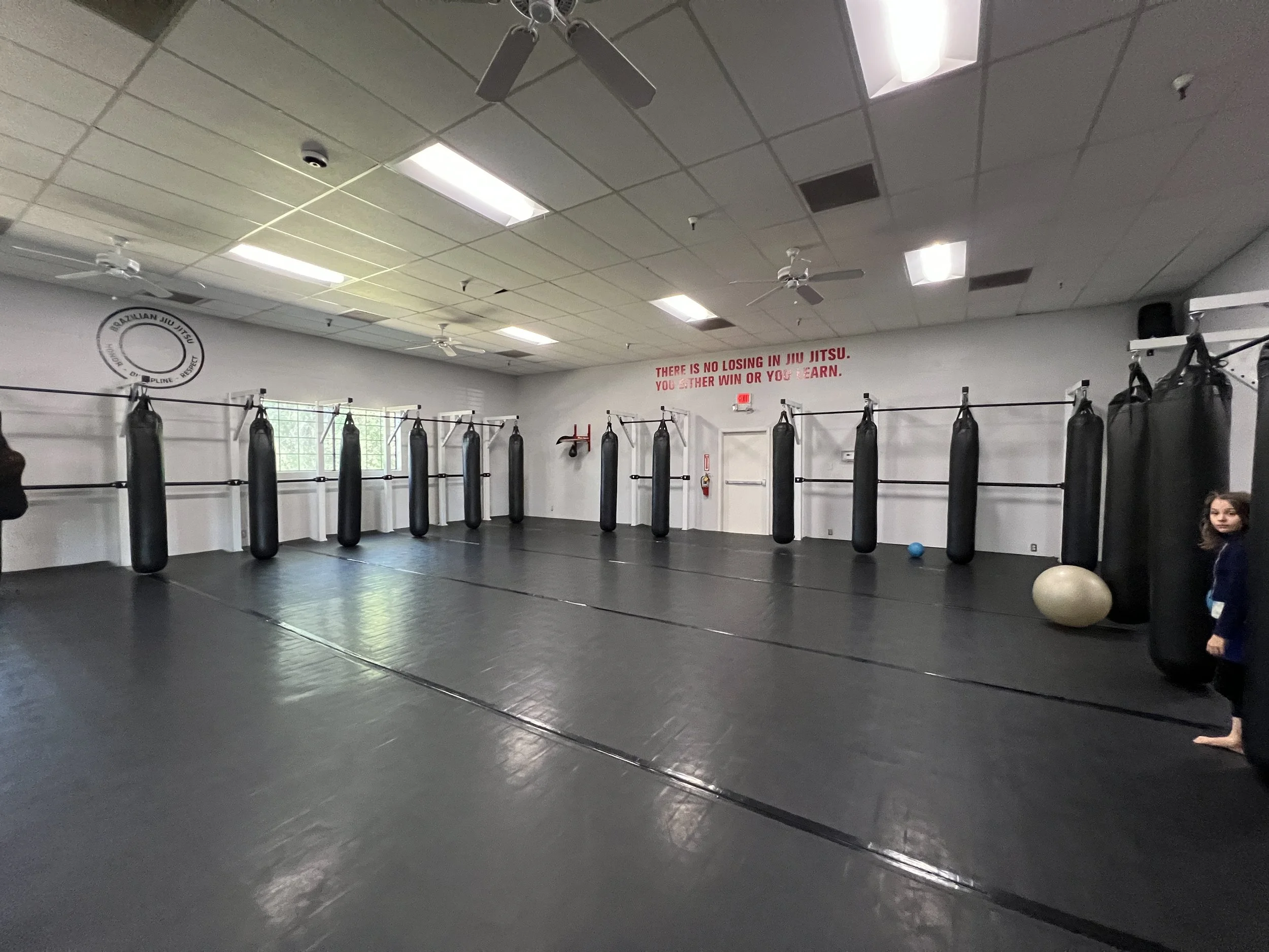 Muay Thai, Boxing, and Brazilian Jiu Jitsu training room with punching bags hanging from the ceiling, black mats on the floor, and motivational text on the wall.