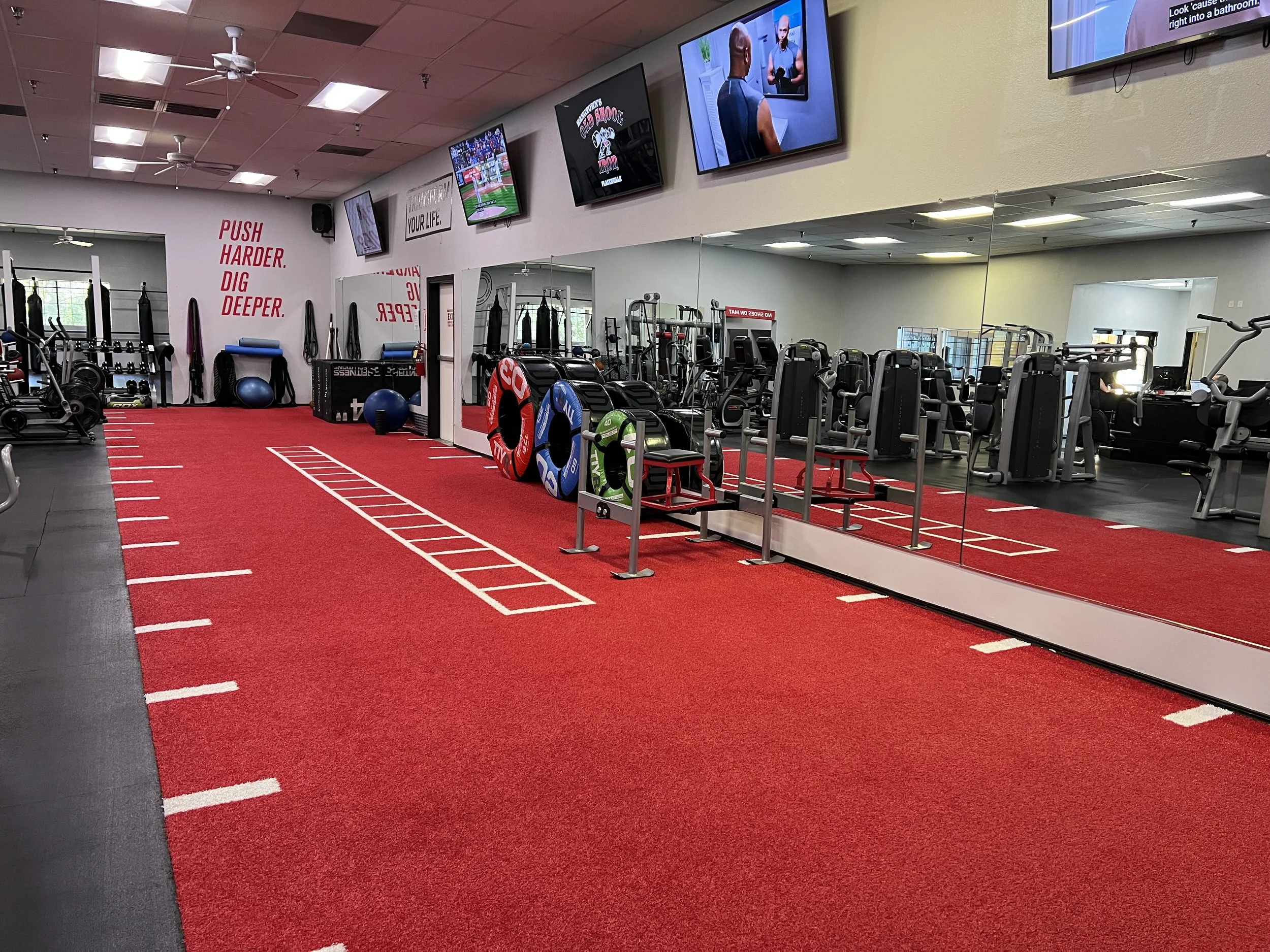 Interior of a gym with a red workout mat on the floor, gym equipment, and multiple television screens on the wall.