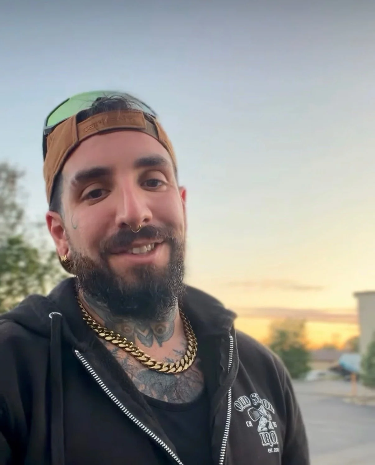 A smiling man with a beard and tattoos on his chest. He is wearing a tan cap backwards, gold chains, earrings, and a black zip-up hoodie with white text. The background shows trees and a partly cloudy sky during sunset.