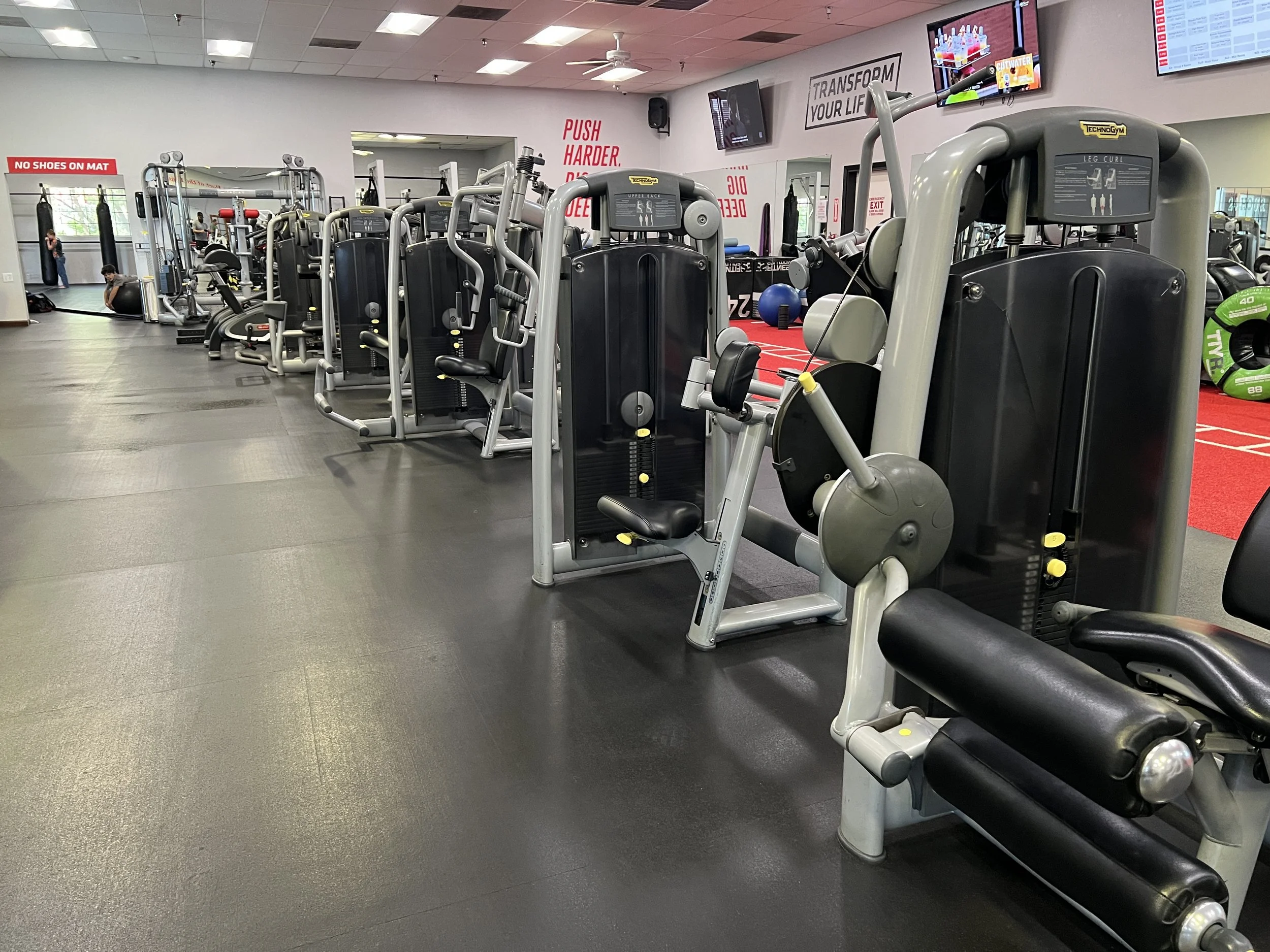 Gym with various workout machines arranged in rows, with a television mounted on the wall and signs that say 'No Shoes on Mat' and 'Push Harder, Deliberate'.