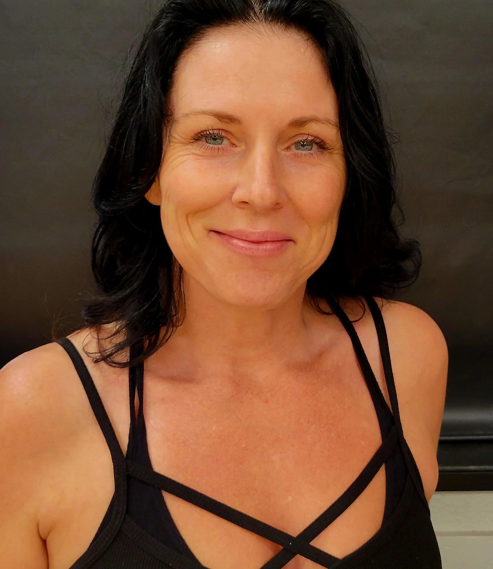 Close-up of a woman with dark curly hair, light skin, wearing a black strappy top, smiling softly, in front of a dark background.