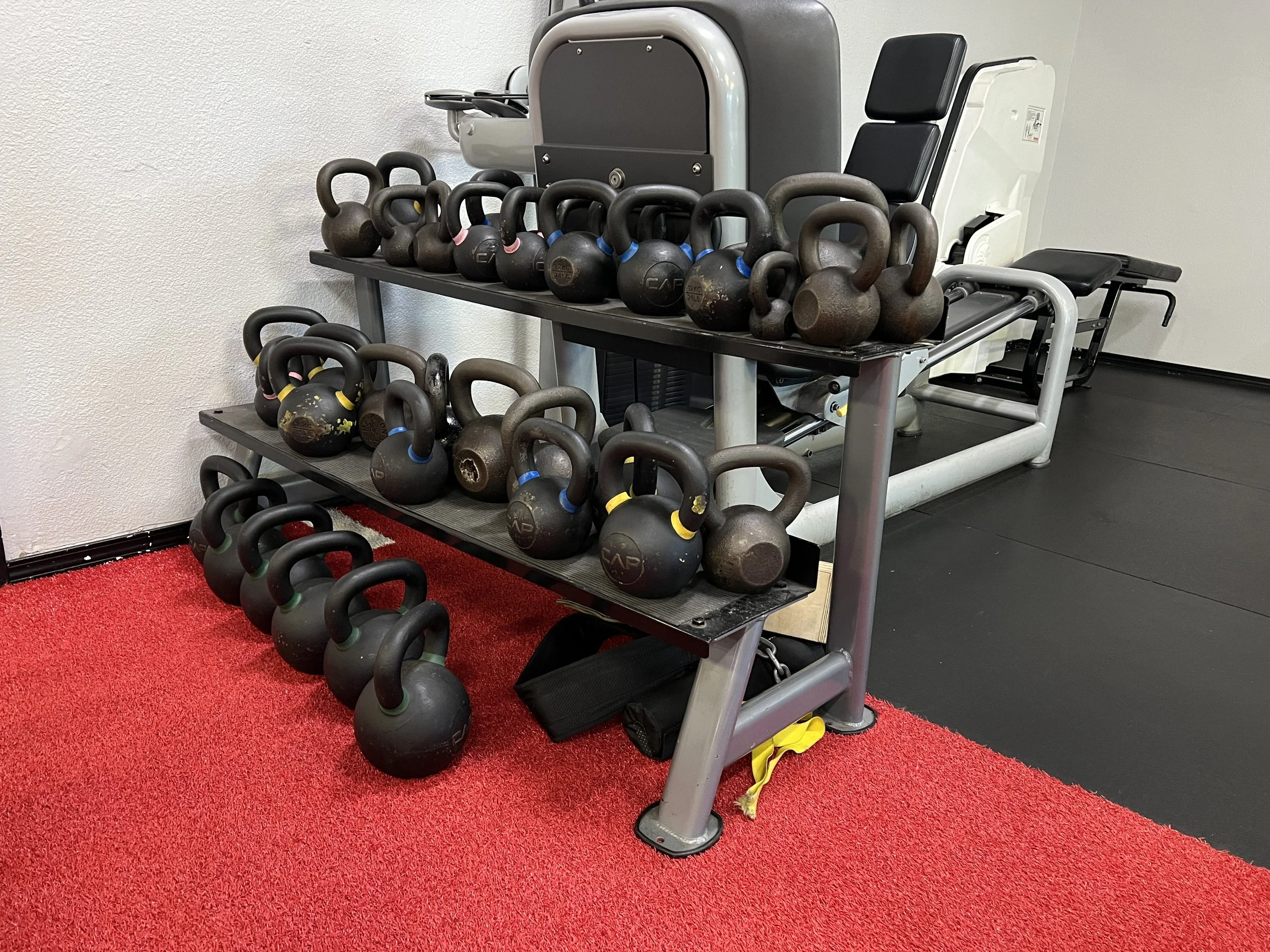 Gym kettlebells placed on a multi-tiered rack, with some on the red carpeted floor nearby, in a fitness center.