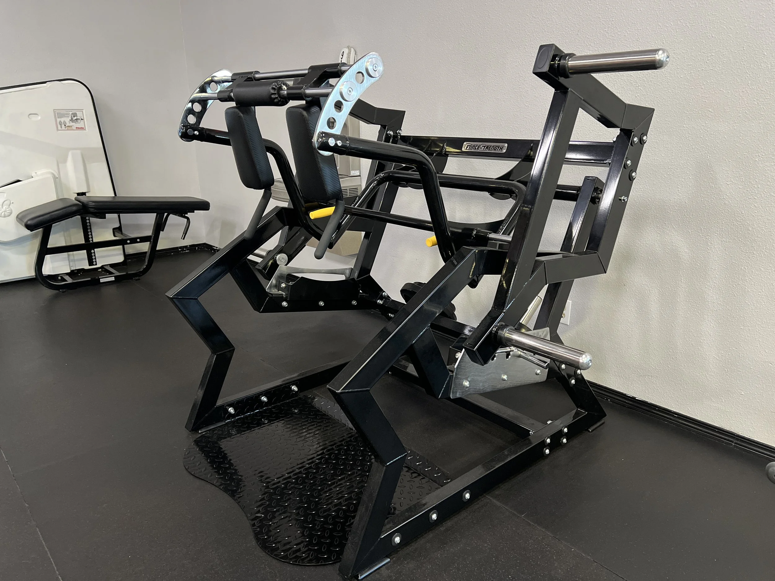 A black leg press exercise machine with padded back and seat, located on a black rubber mat in a gym setting with plain white walls.