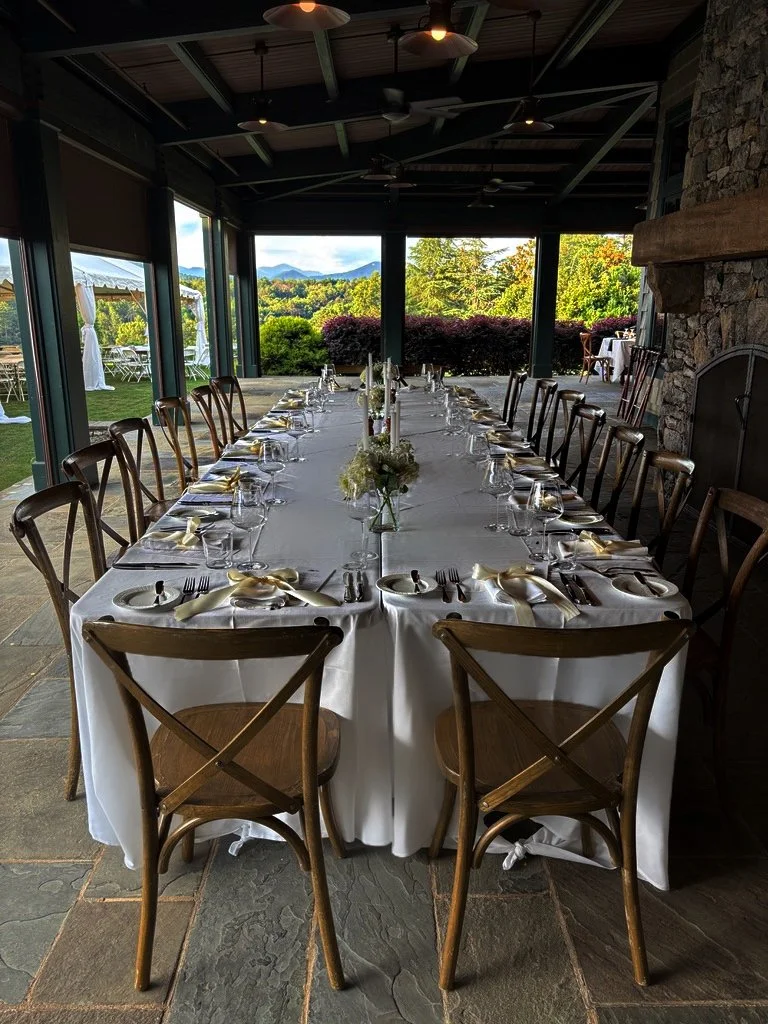 Long indoor dining table set for a meal with white tablecloths, floral centerpieces, and place settings including plates, silverware, and glasses, in a room with large windows showing outdoor scenery.