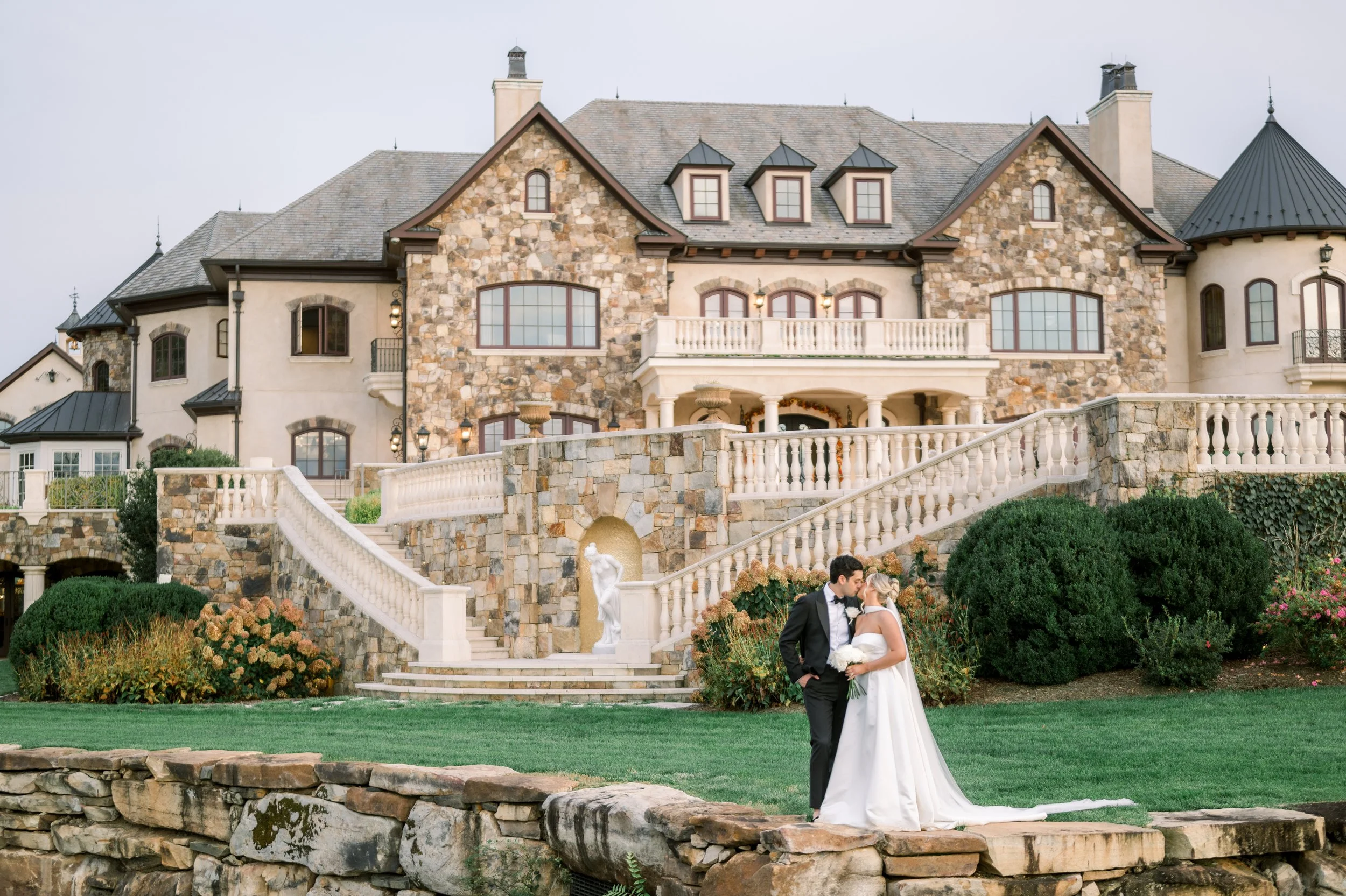 A bride and groom stand in front of a large, stone and stucco mansion with multiple turrets and gabled roofs, during a wedding celebration outside.