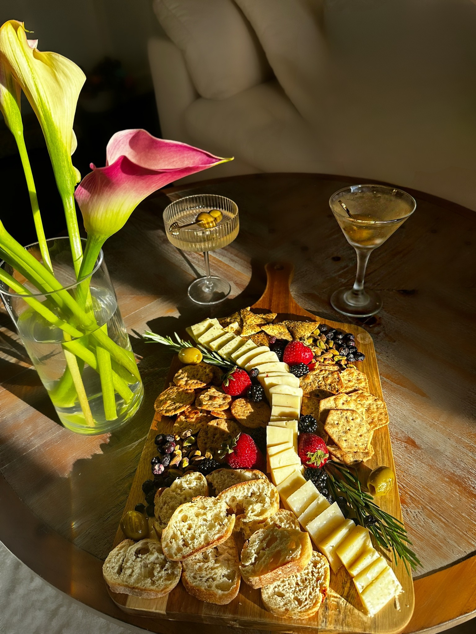 A wooden cheese platter with various cheeses, strawberries, blackberries, nuts, and crackers. In the background, there is a bouquet of pink and yellow lilies in a glass vase, a martini glass with a drink, and a small dish with an olive. The setting appears to be on a round wooden table near a beige couch.