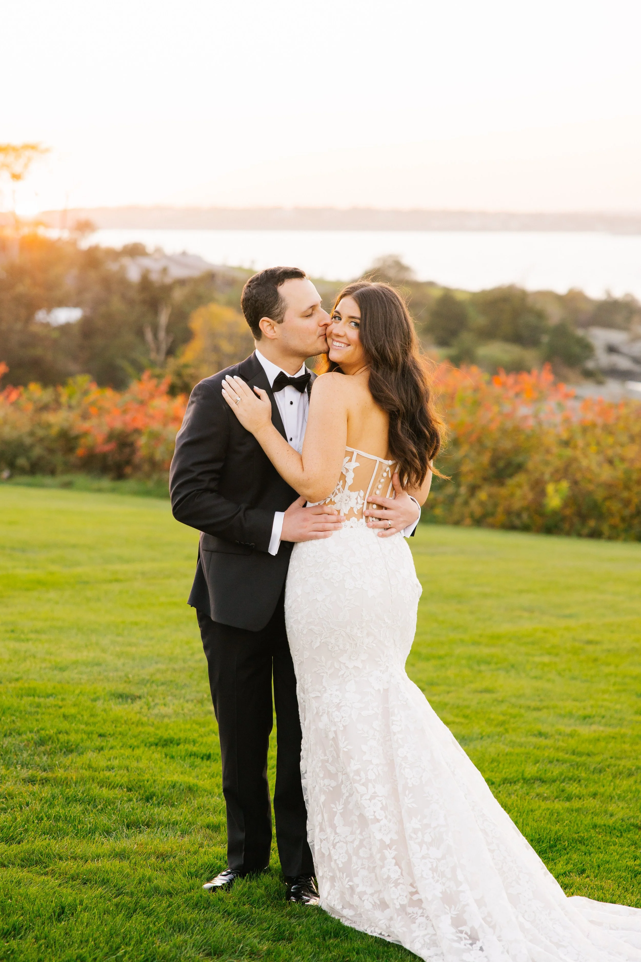 A newlywed couple stands outdoors on a grassy field, with the groom kissing the bride on the cheek and the bride smiling at the camera, during sunset with colorful trees in the background.