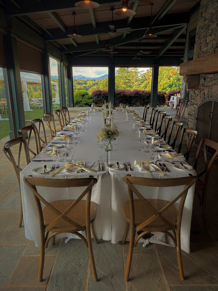 Restaurant dining area with a long table set with white tablecloth, napkins, glassware, and floral centerpieces, overlooking a lush outdoor landscape with mountains.
